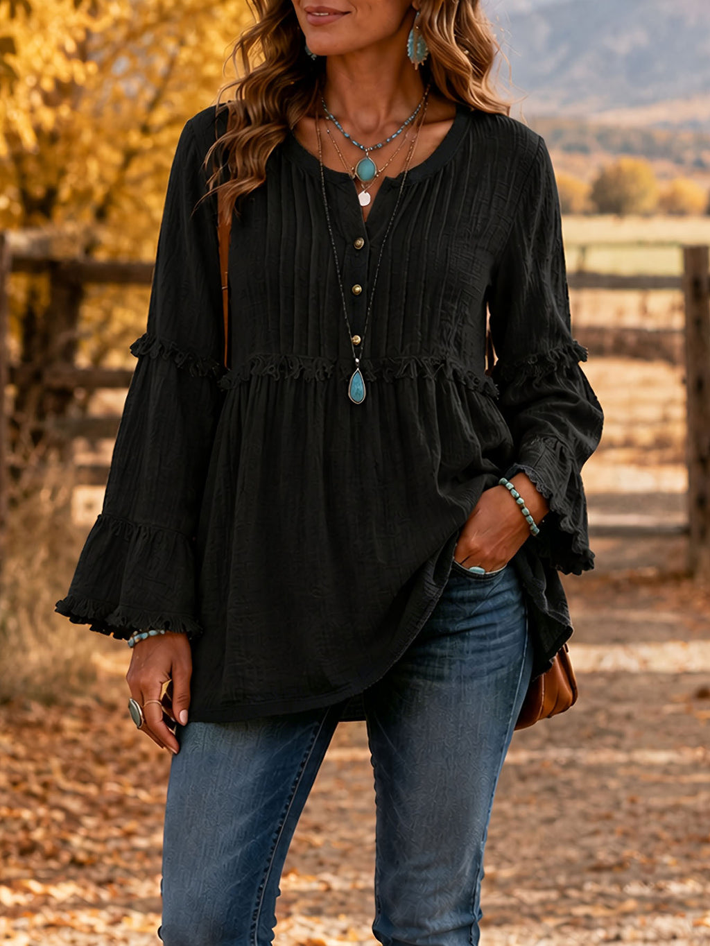 A woman wearing a black long-sleeve blouse with pleated details and ruffled cuffs, paired with blue jeans, standing outdoors on a path with autumn trees in the background.