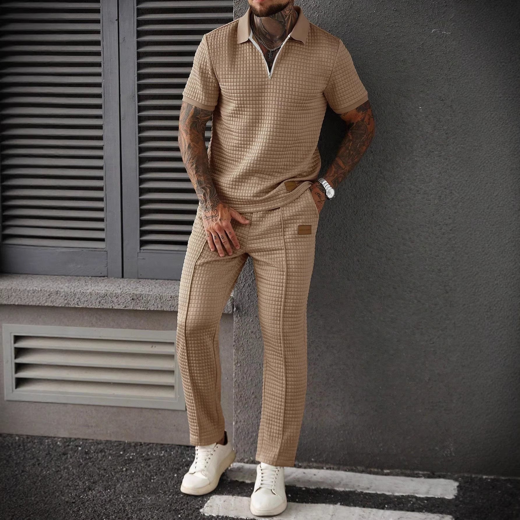 Man wearing a beige checkered outfit standing against a gray wall.