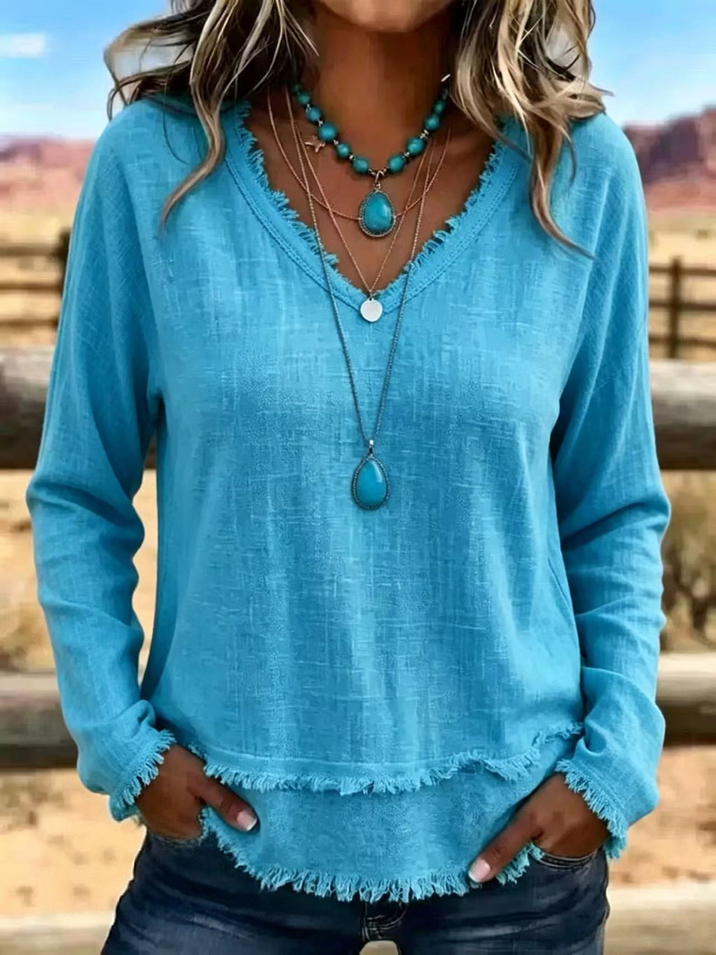 A woman wearing a turquoise blue long-sleeve top with a frayed hem and neckline, layered with turquoise and silver necklaces, standing outdoors in a desert landscape.