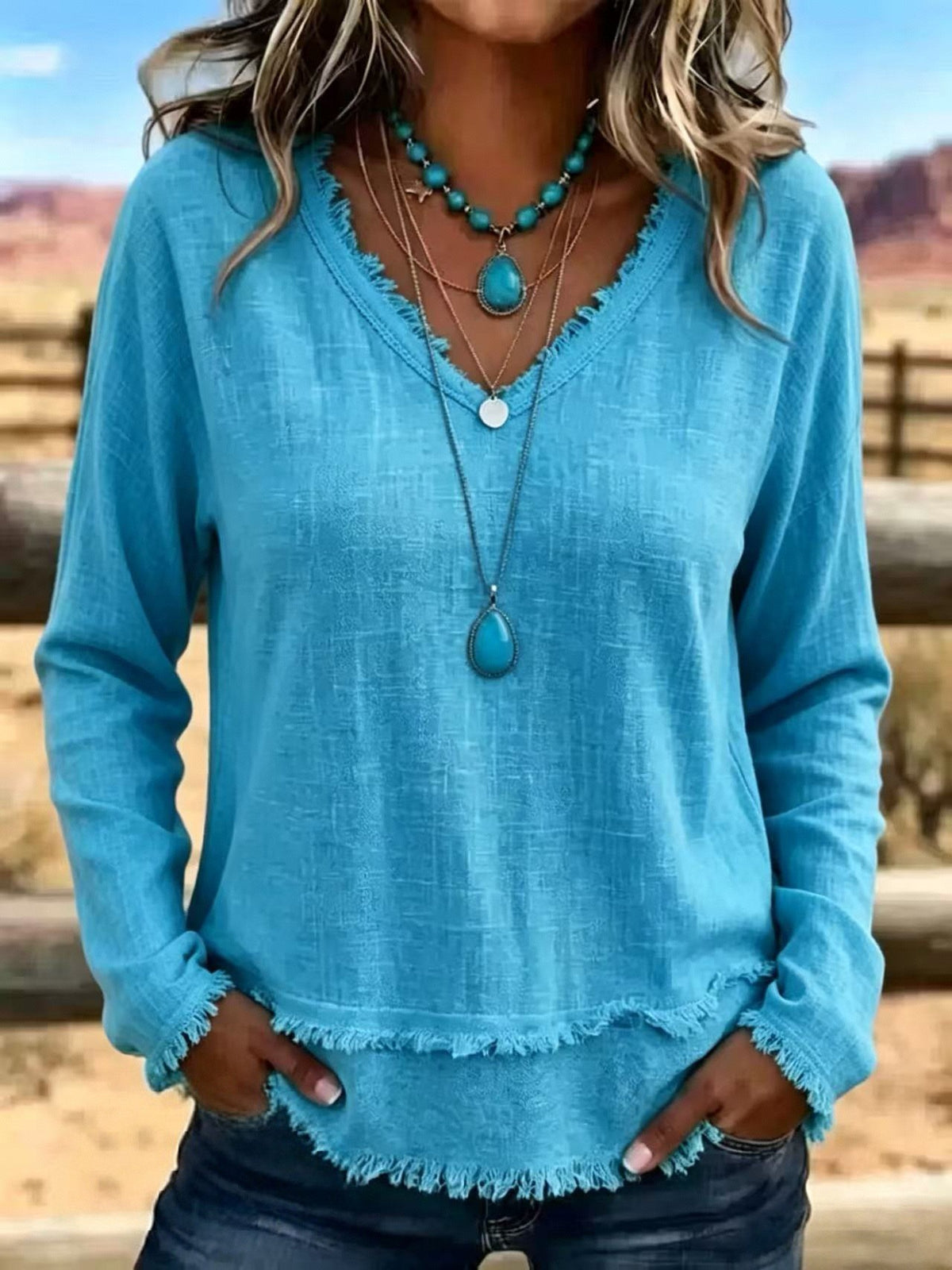 A woman wearing a turquoise blue long-sleeve top with a frayed hem and neckline, layered with turquoise and silver necklaces, standing outdoors in a desert landscape.