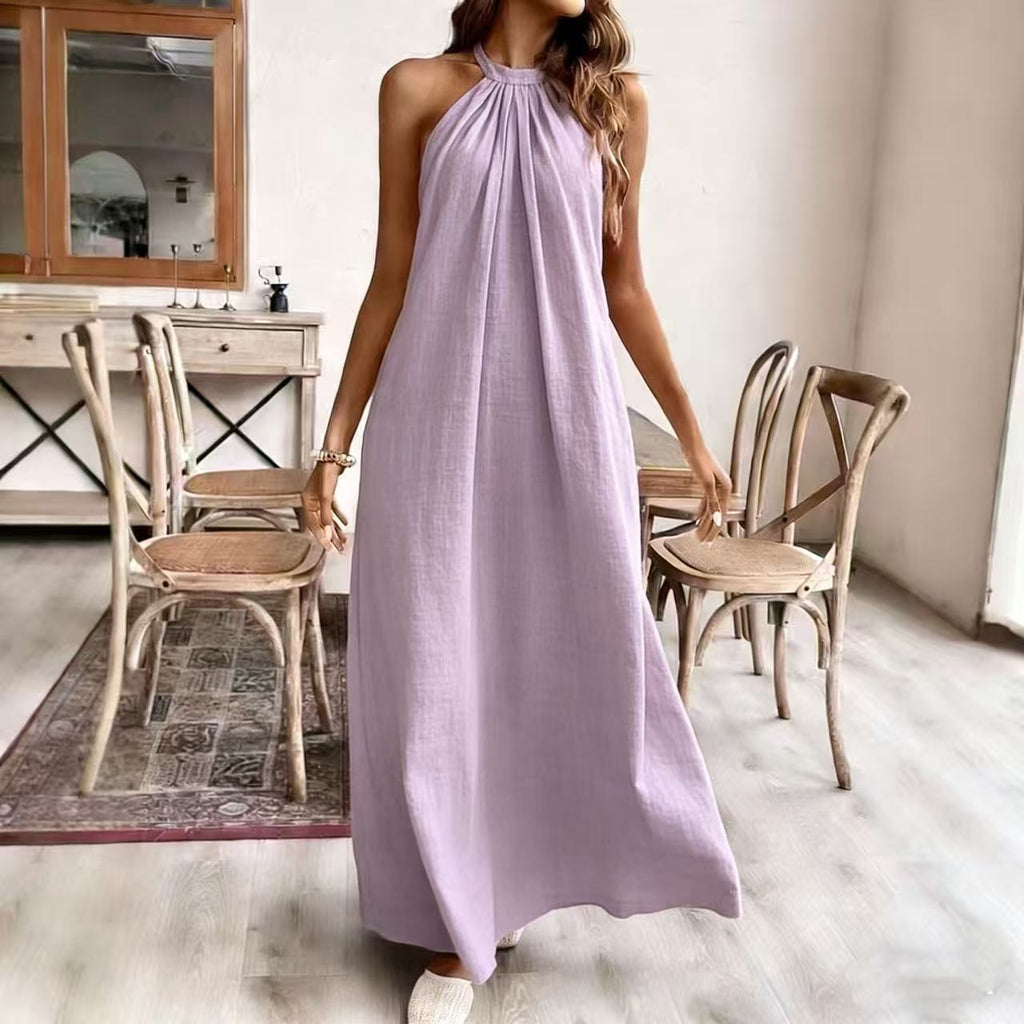 Woman in a long lilac dress standing in a room with wooden chairs and a mirror.