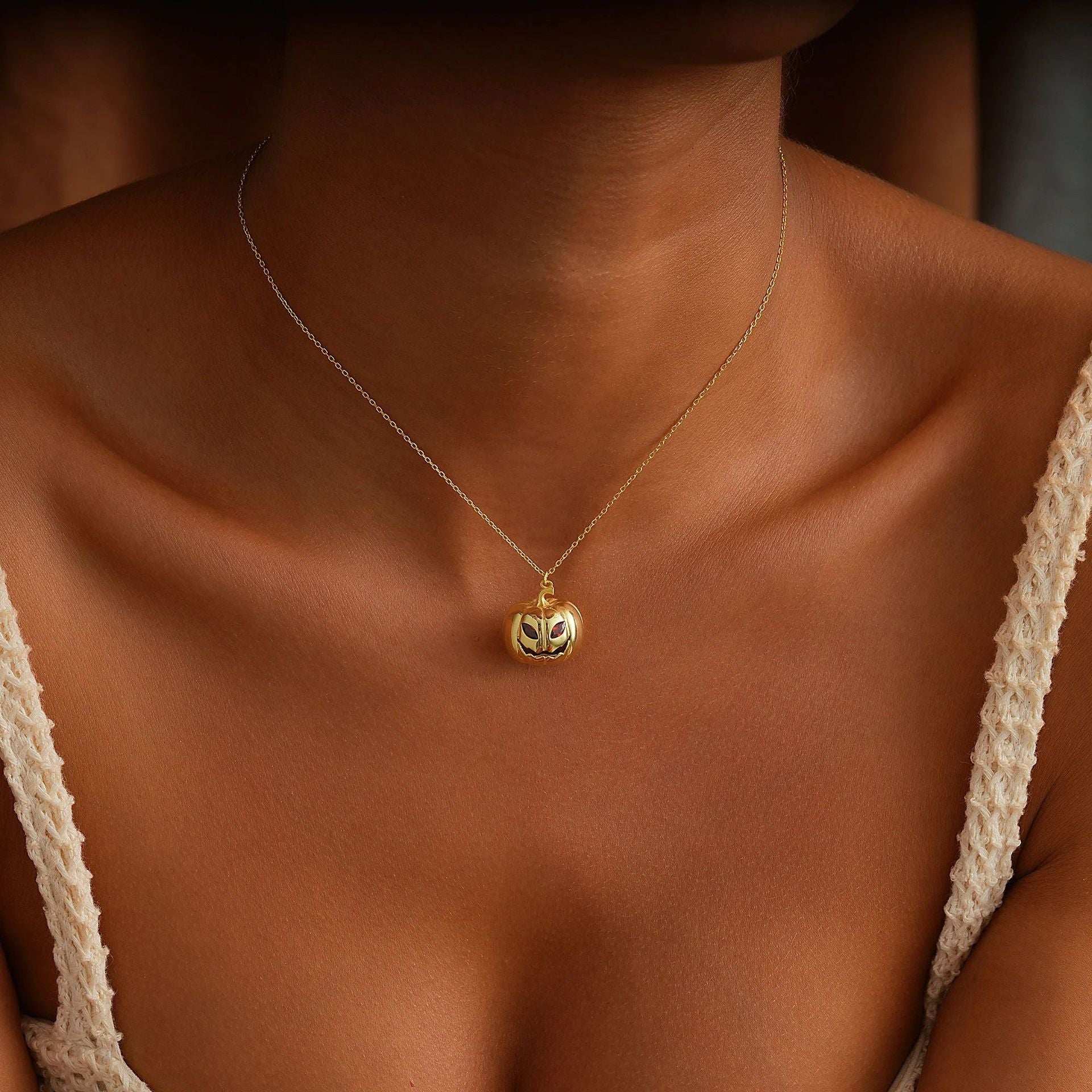 A close-up of a model's neck and chest wearing a delicate gold chain necklace with a small round gold pendant featuring a stylized face.