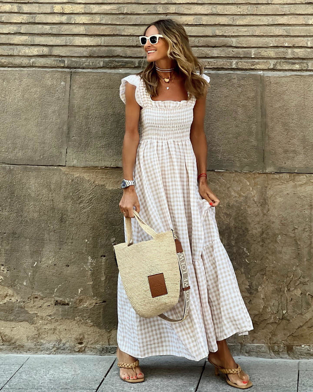 Woman in a white dress holding a beige bag against a stone wall.