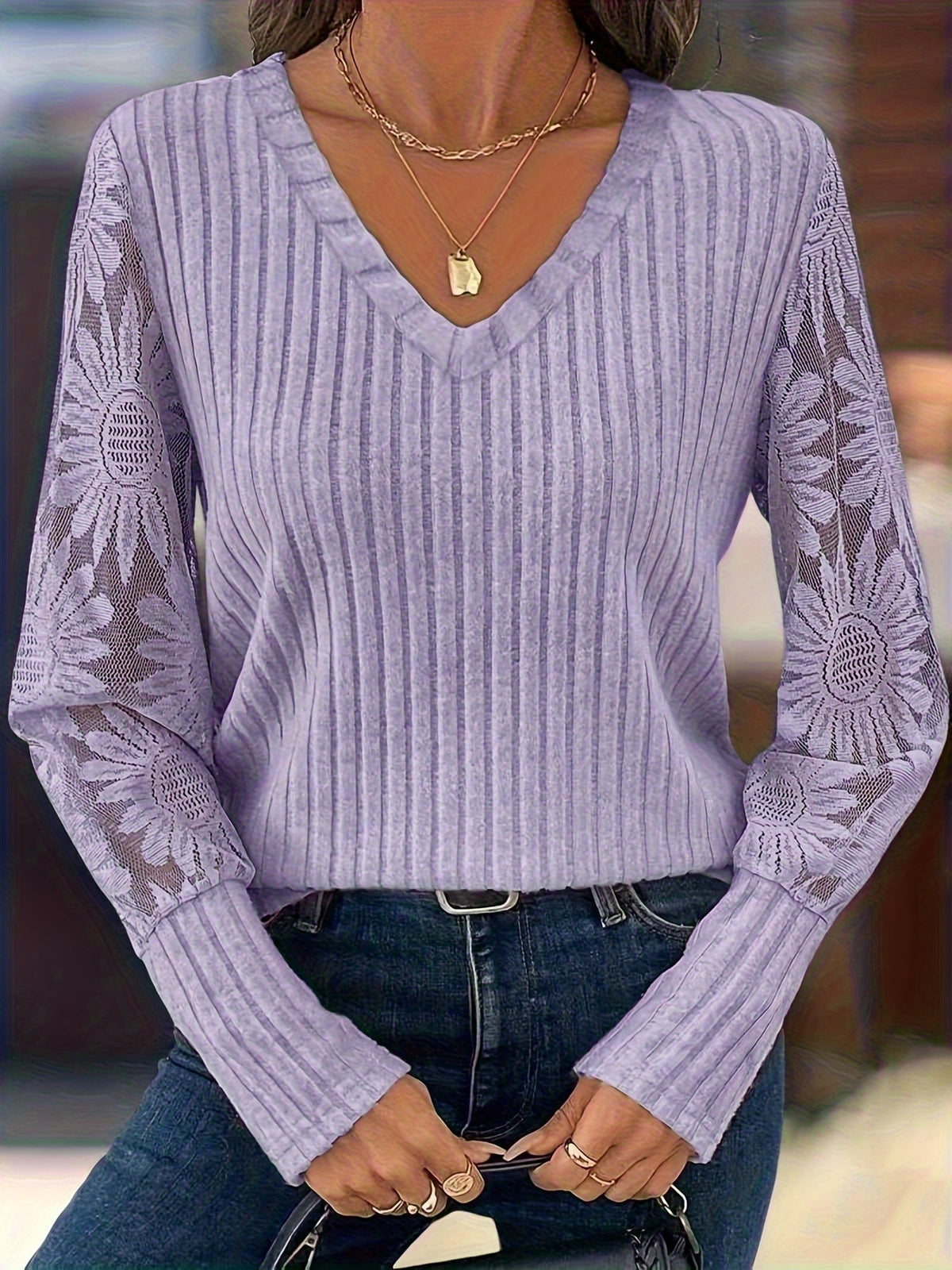 A woman wearing a lavender ribbed knit sweater with floral lace sleeves, paired with dark blue jeans and layered gold necklaces.