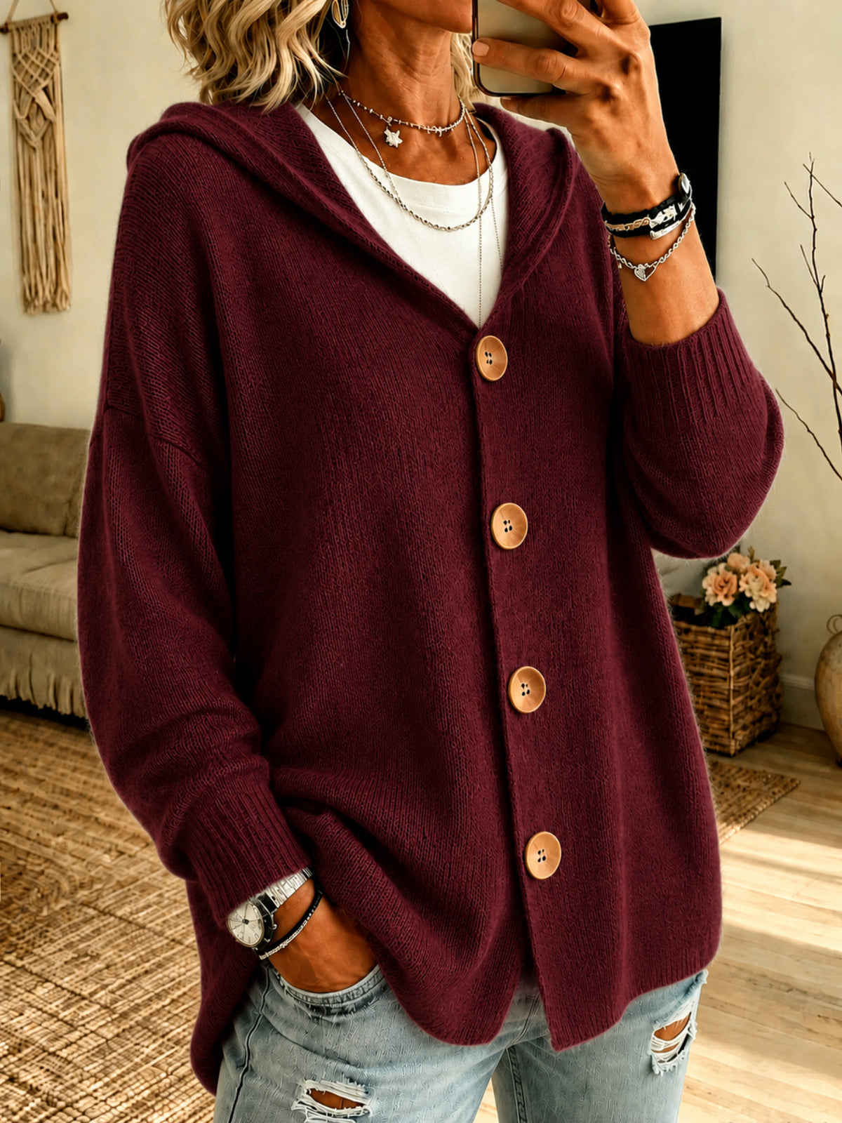A woman wearing an oversized maroon knit cardigan with a hood and large wooden buttons, layered over a white top and paired with distressed light blue jeans in a cozy indoor setting.