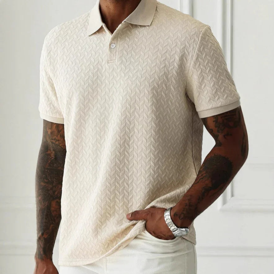 Man wearing a beige patterned polo shirt with tattoos on arms against a white background