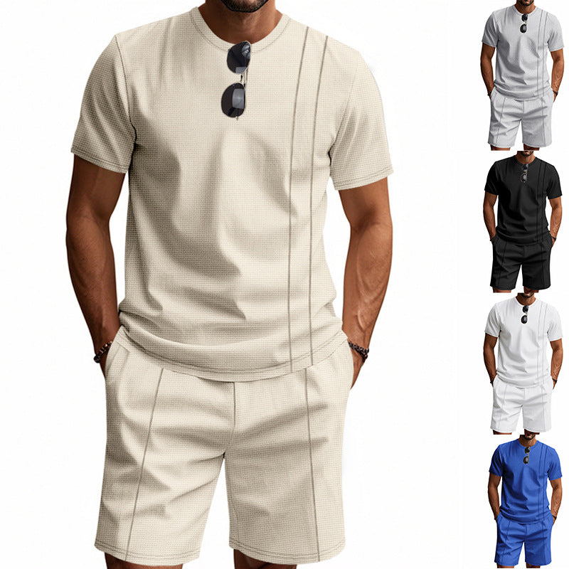 Man wearing a beige short-sleeve shirt and shorts with sunglasses on his shirt, against a white background.