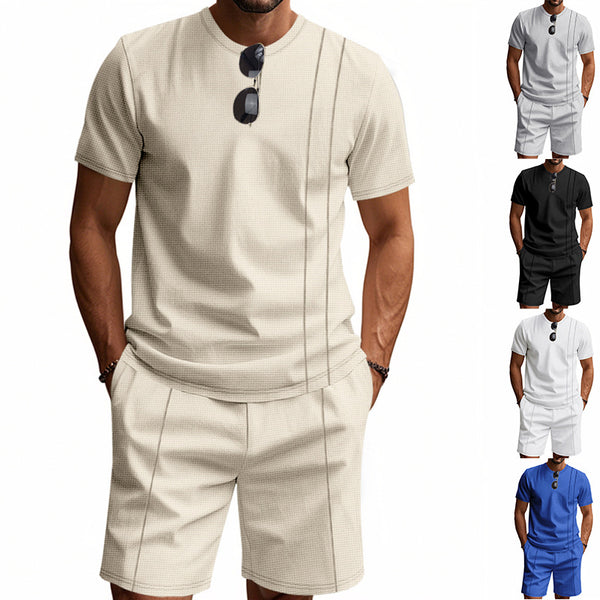 Man wearing a beige short-sleeve shirt and shorts with sunglasses on his shirt, against a white background.