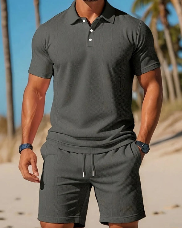 Man wearing a gray polo shirt and shorts set on a beach.