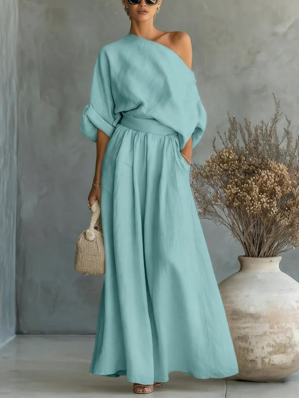 Woman wearing a light blue dress standing next to a vase with dried flowers against a gray wall.