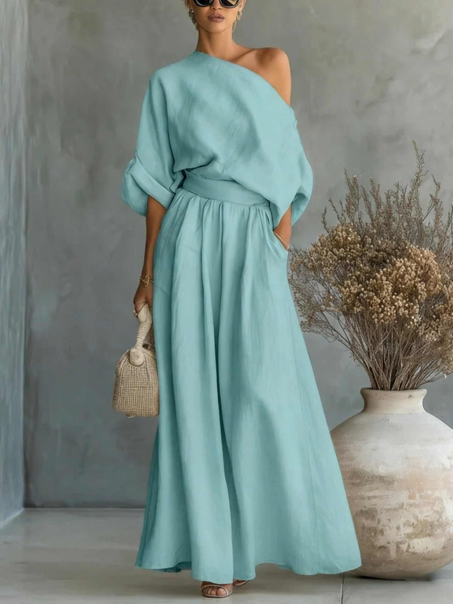 Woman wearing a light blue dress standing next to a vase with dried flowers against a gray wall.