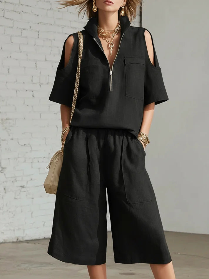 Person wearing a black outfit with cut-out shoulders against a white brick wall.