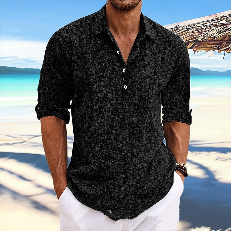Man wearing a black button-up shirt and white pants on a beach with a clear blue sky.