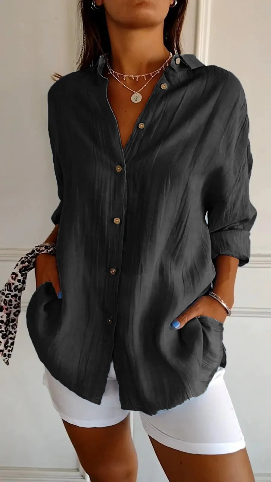 A woman wearing a dark charcoal gray crinkled button-down shirt with rolled-up sleeves and white shorts, accessorized with layered gold necklaces.