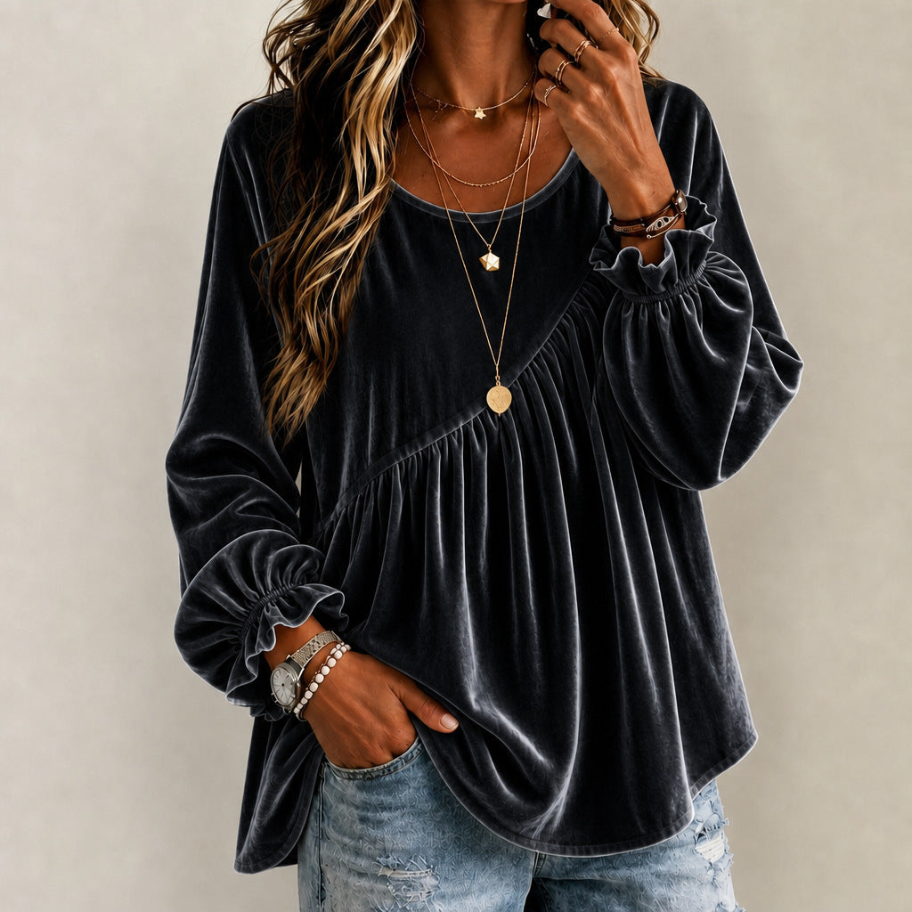 A woman wearing a black velvet long-sleeve blouse with ruffled cuffs and a gathered peplum-style waist, paired with light blue jeans and layered gold necklaces, against a neutral beige background.
