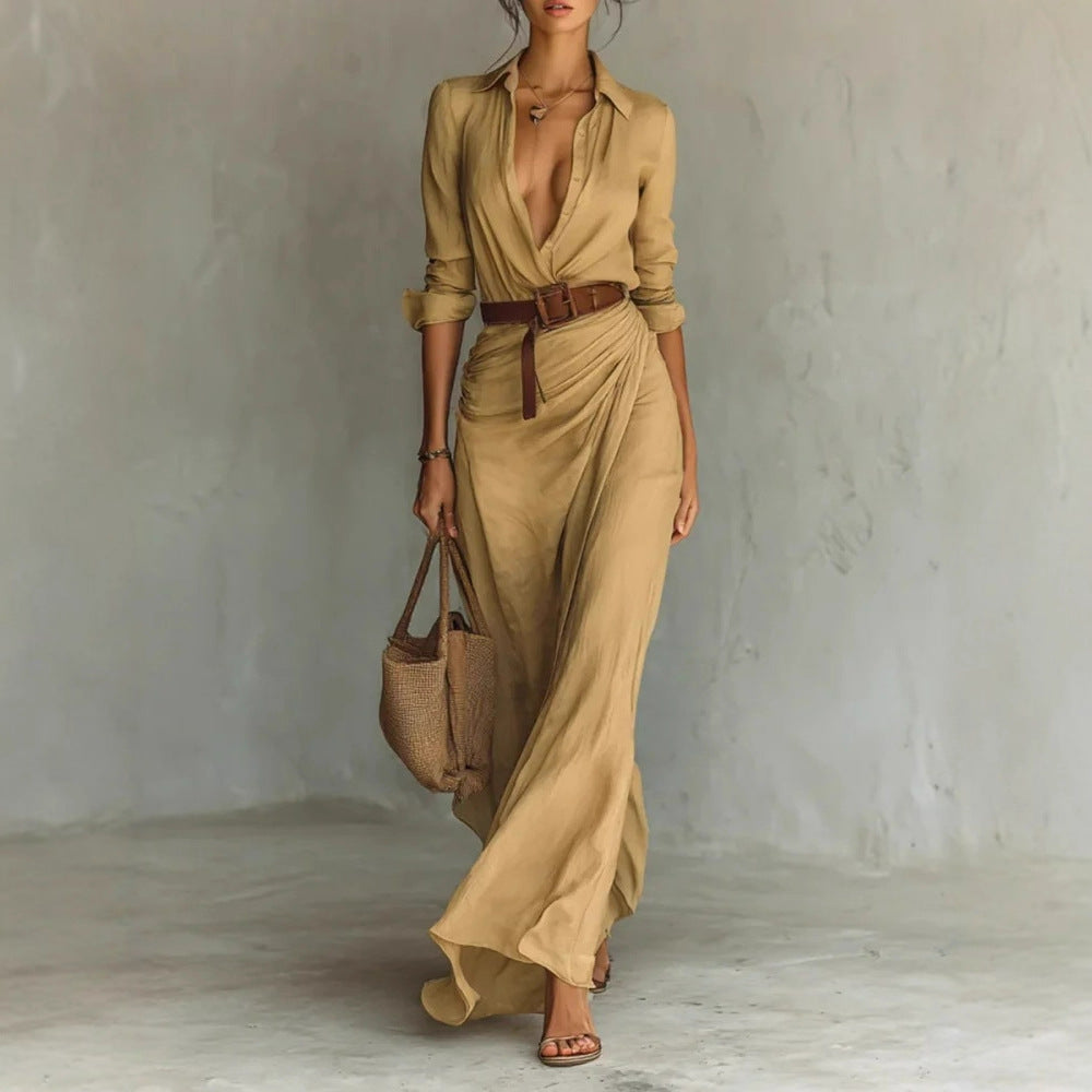 Woman wearing a long beige dress with a belt, holding a woven bag against a plain background.
