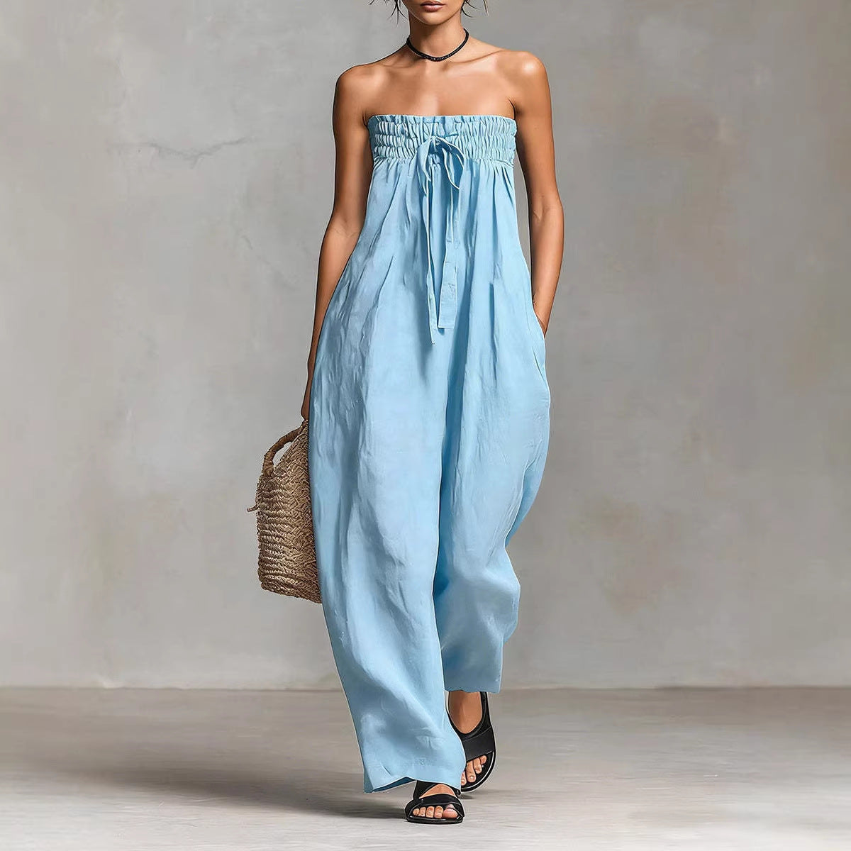 Woman wearing a light blue strapless jumpsuit holding a woven bag against a neutral background