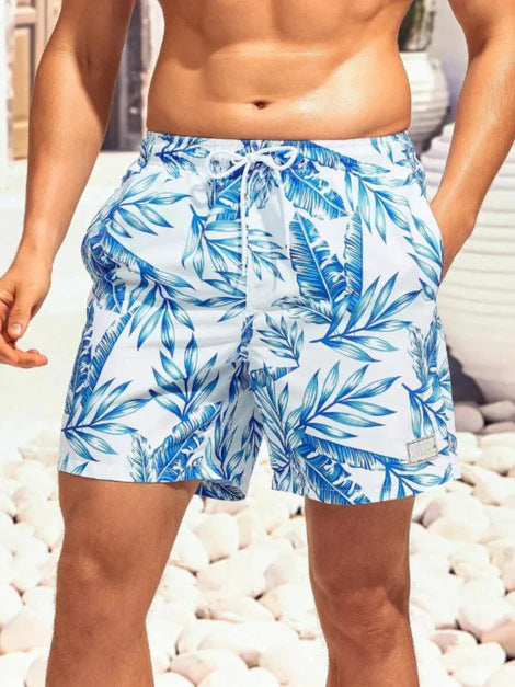 Person wearing blue tropical leaf patterned shorts on a pebble beach.