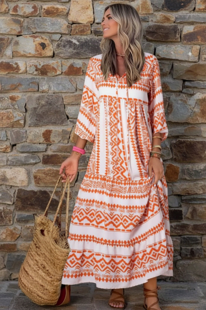 Woman wearing an orange and white patterned dress holding a woven bag standing against a stone wall.