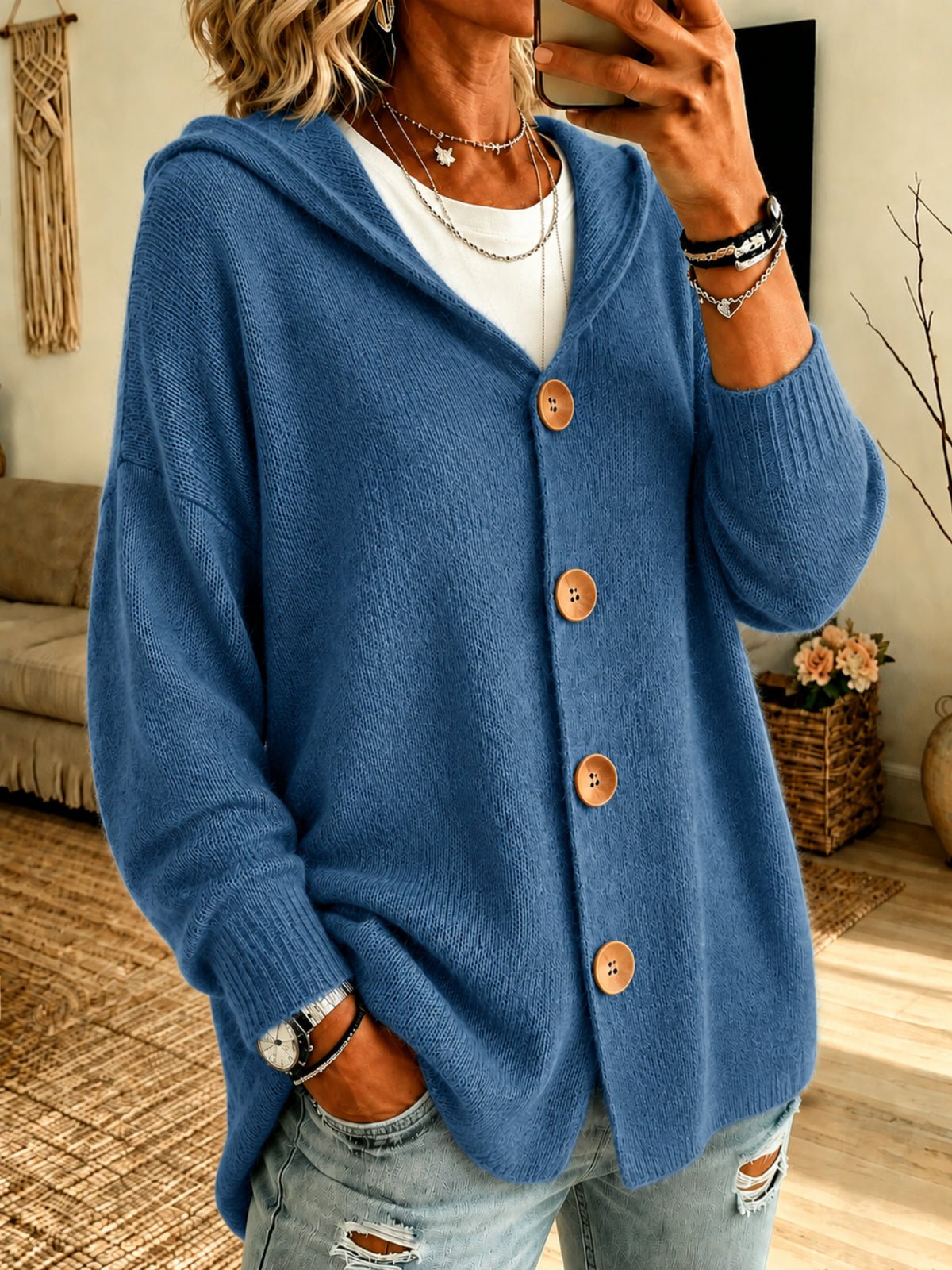 A woman wearing a blue knit hooded cardigan with large brown buttons over a white top and light blue ripped jeans, photographed in a home interior setting.