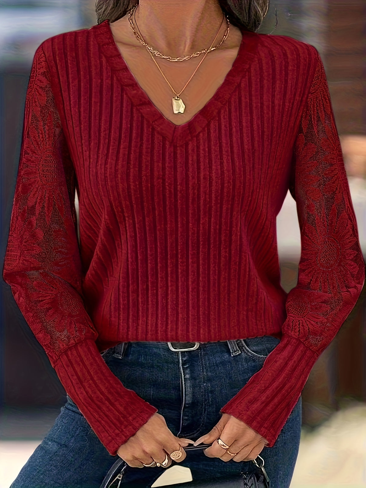 A woman wearing a long-sleeve ribbed knit sweater in deep red, featuring sheer lace-like sleeves and a V-neckline, paired with blue jeans.