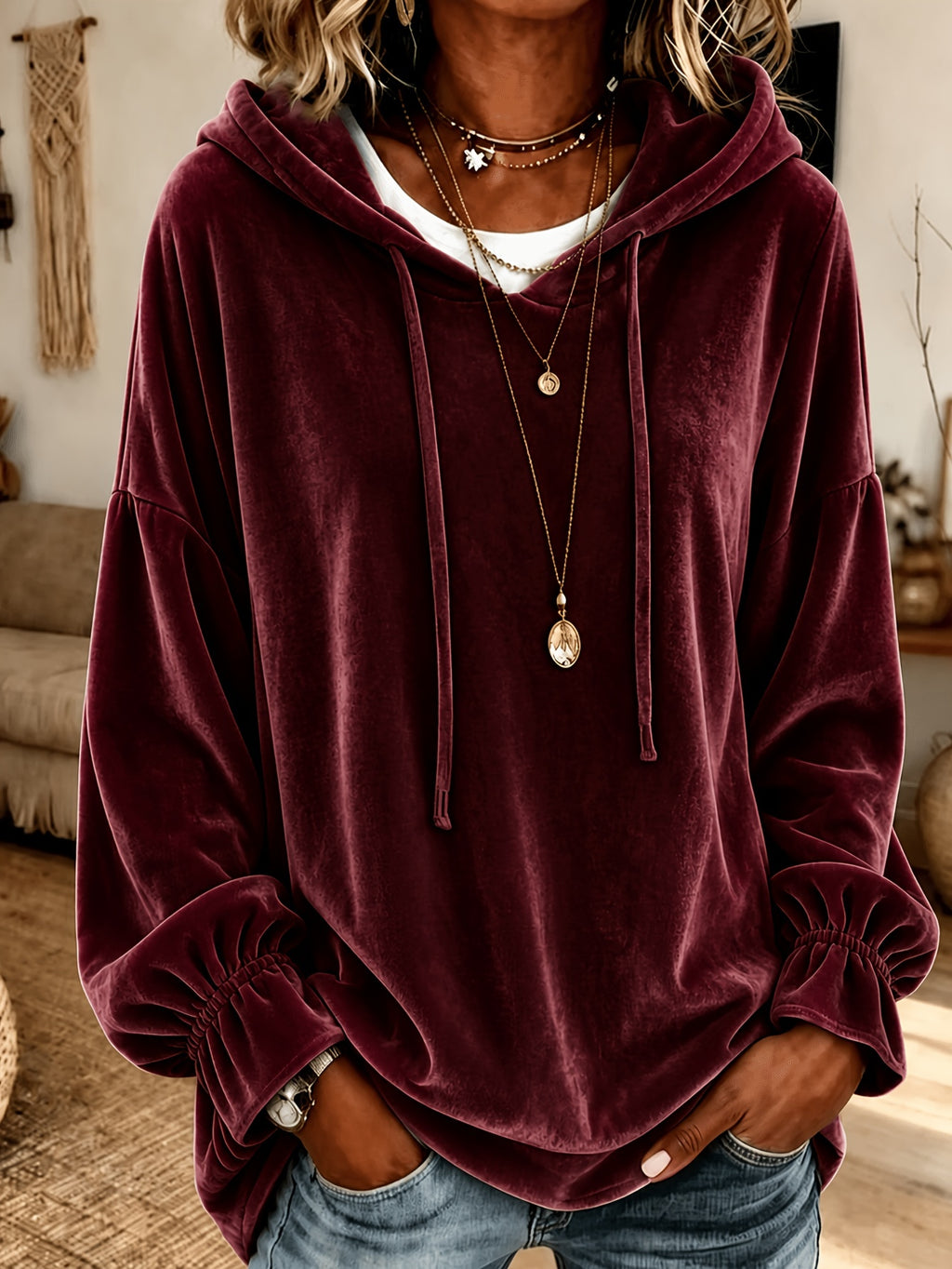 A woman wearing a burgundy velvet pullover hoodie with drawstrings and elasticized cuffs, layered over a white t-shirt and blue jeans in a cozy indoor setting.