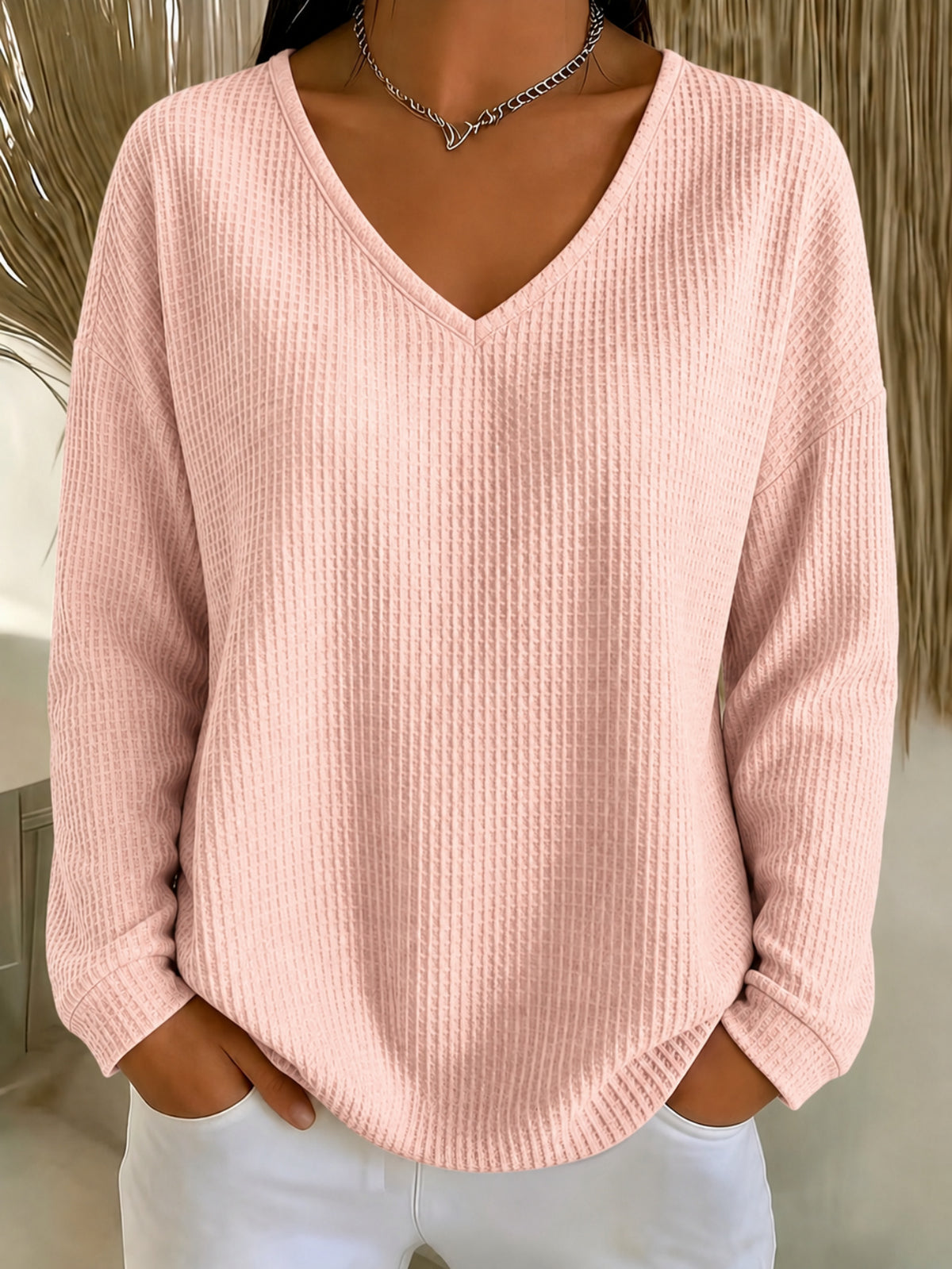 A woman wearing a light pink waffle-knit long-sleeve sweater with a V-neck, paired with white pants and a silver necklace.