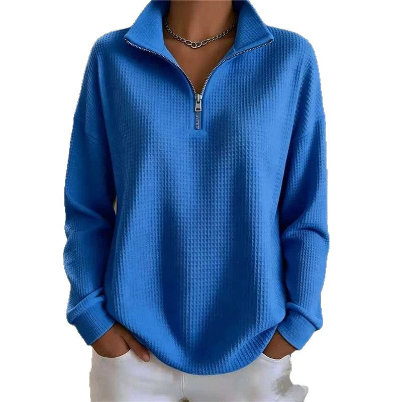 A woman wearing a bright blue waffle-knit quarter-zip pullover sweater with a stand collar, paired with white pants and a silver chain necklace, shown against a plain white background.