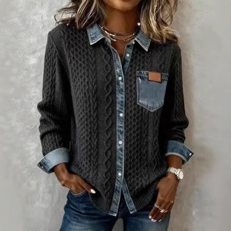 Person wearing a dark cable knit sweater over a denim shirt with rolled-up sleeves against a neutral background.