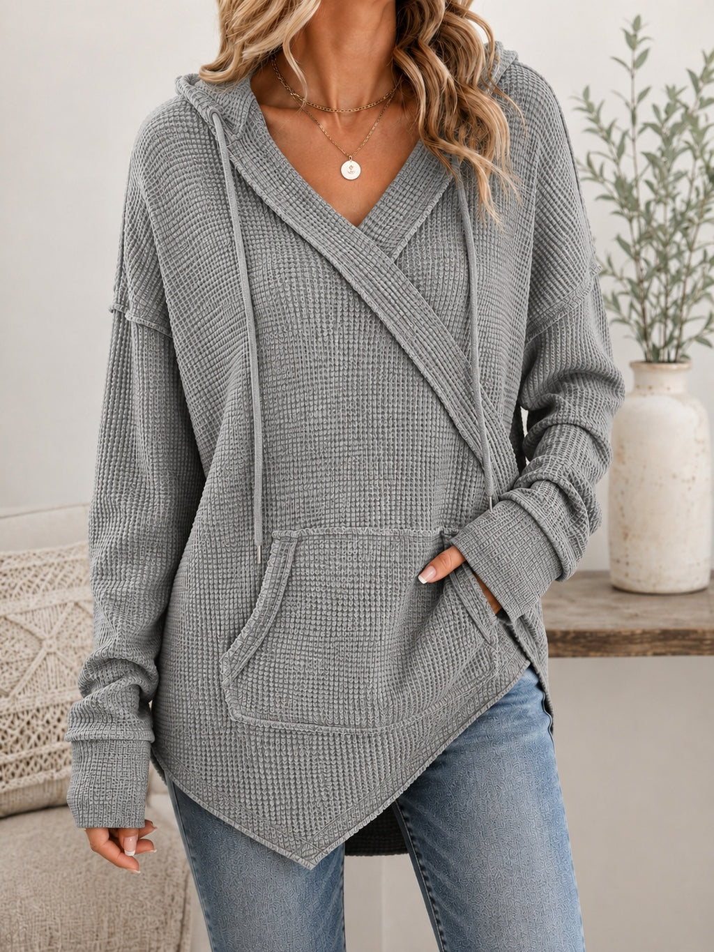 A woman wearing a gray waffle-knit hooded wrap sweater with drawstrings and front pockets, paired with blue jeans, in a softly styled indoor setting.