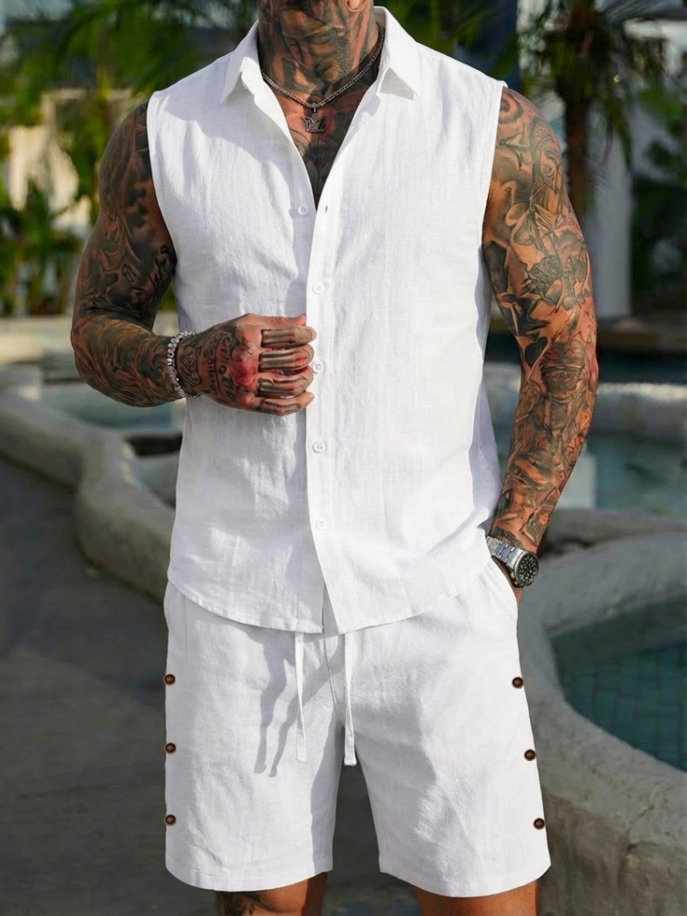 Man wearing a white sleeveless shirt and shorts with tattoos, standing by a pool.