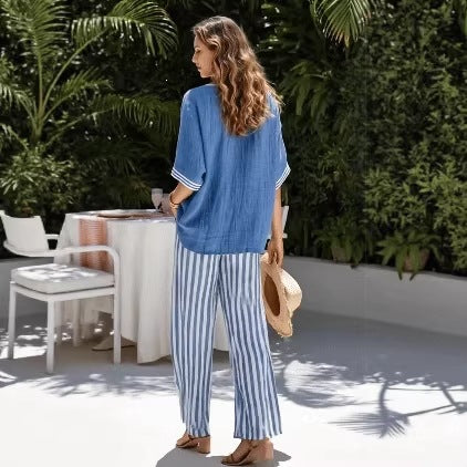 Woman in a blue top and striped pants standing outdoors with greenery and a table in the background.