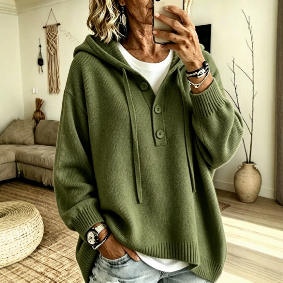 A woman takes a mirror selfie wearing an oversized olive green knit hoodie sweater with a buttoned placket over a white t-shirt and light-wash jeans, in a cozy living room setting.