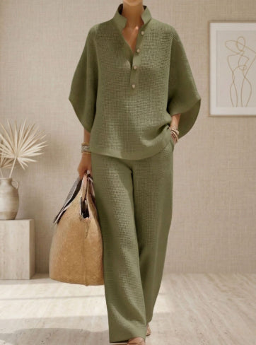 Person wearing a green textured outfit holding a beige bag in a room with a plant and abstract art on the wall.