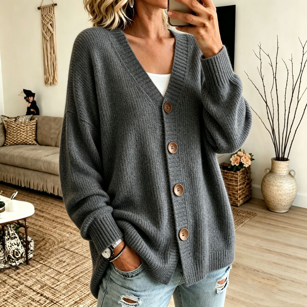 A woman wearing a dark gray chunky knit cardigan with brown buttons over a white top and light blue ripped jeans, standing in a cozy living room setting.