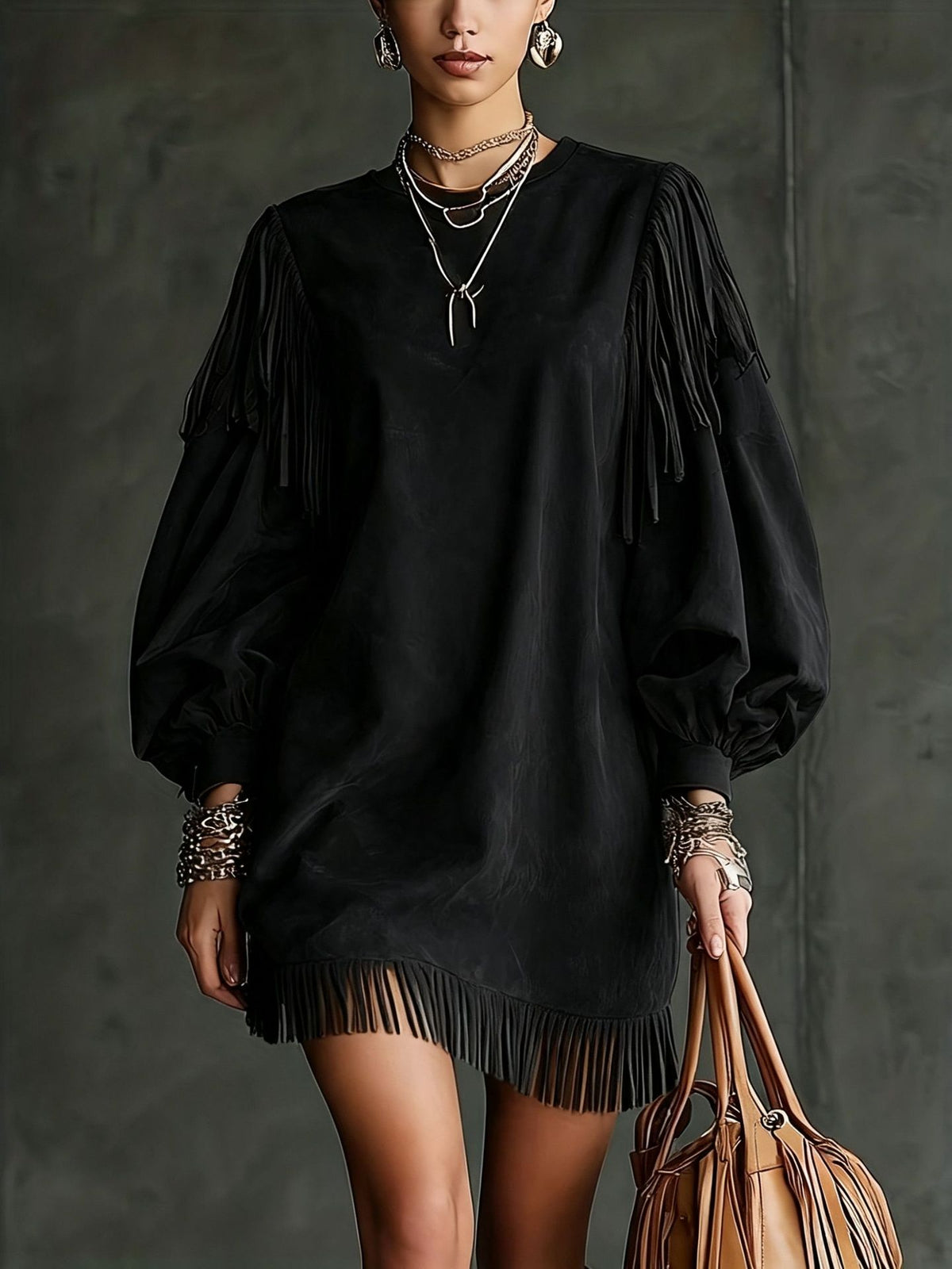 A model wearing a black long-sleeve mini dress with dramatic fringe detailing on the sleeves and hem, accessorized with layered silver necklaces, bracelets, and earrings, while holding a brown leather shoulder bag against a dark gray textured studio background.