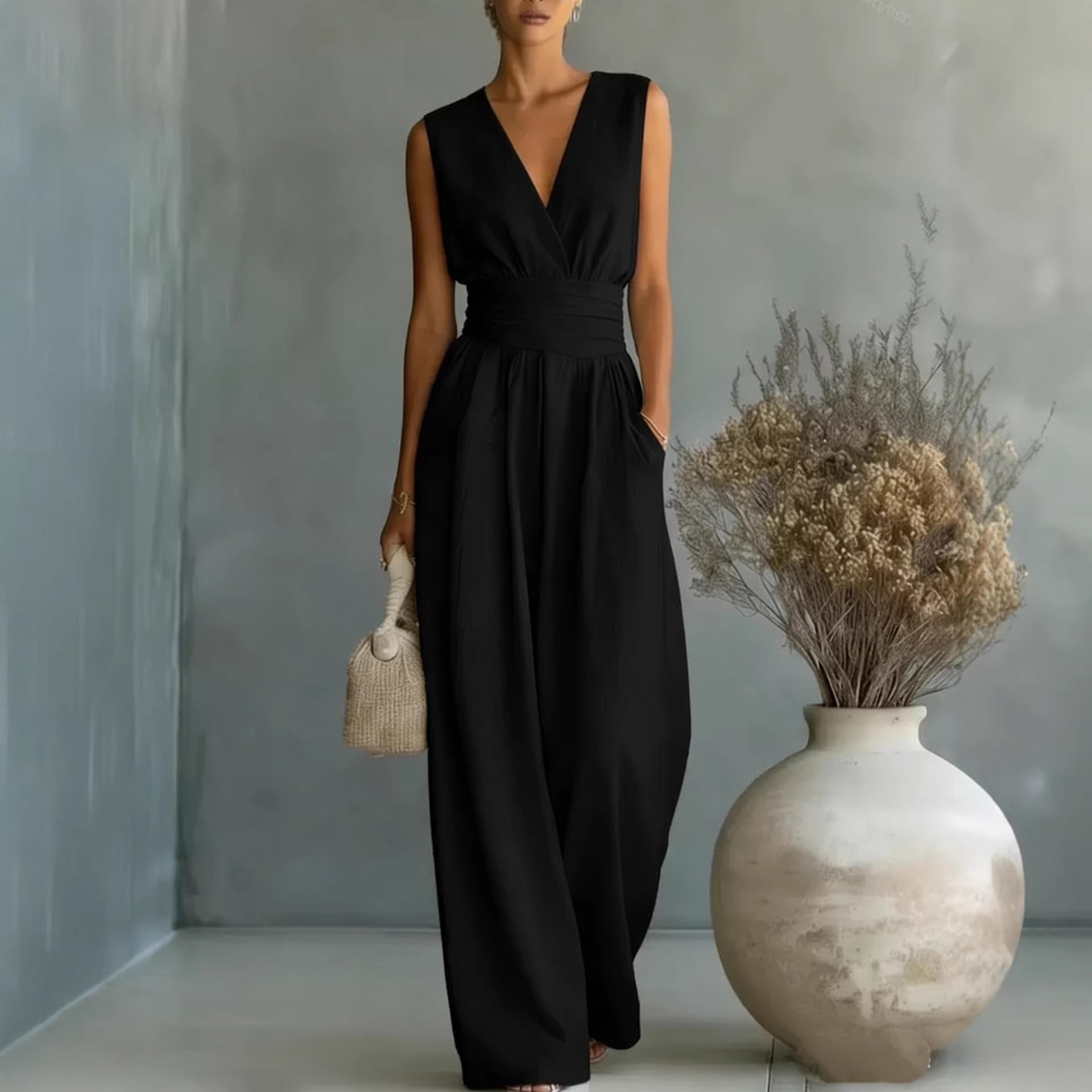 Woman in a black sleeveless jumpsuit standing next to a vase with dried flowers against a gray wall.