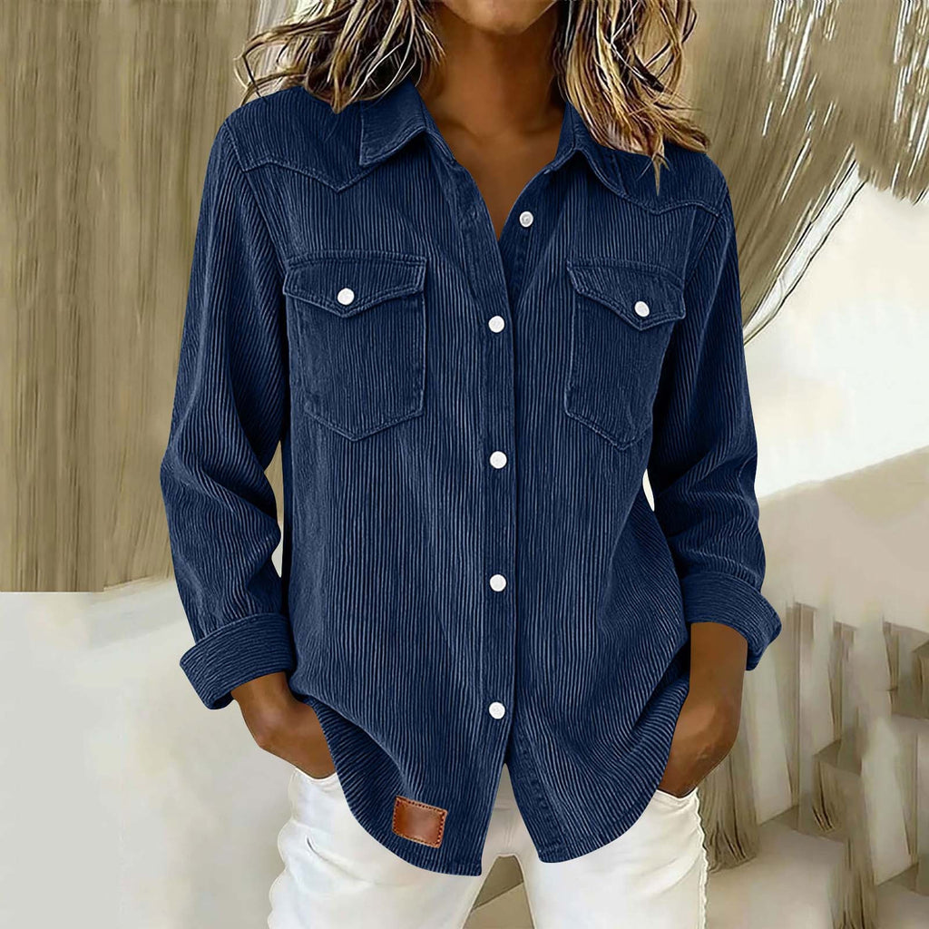 A woman wearing a dark blue corduroy button-up shirt with two chest pockets and white pants, standing indoors.