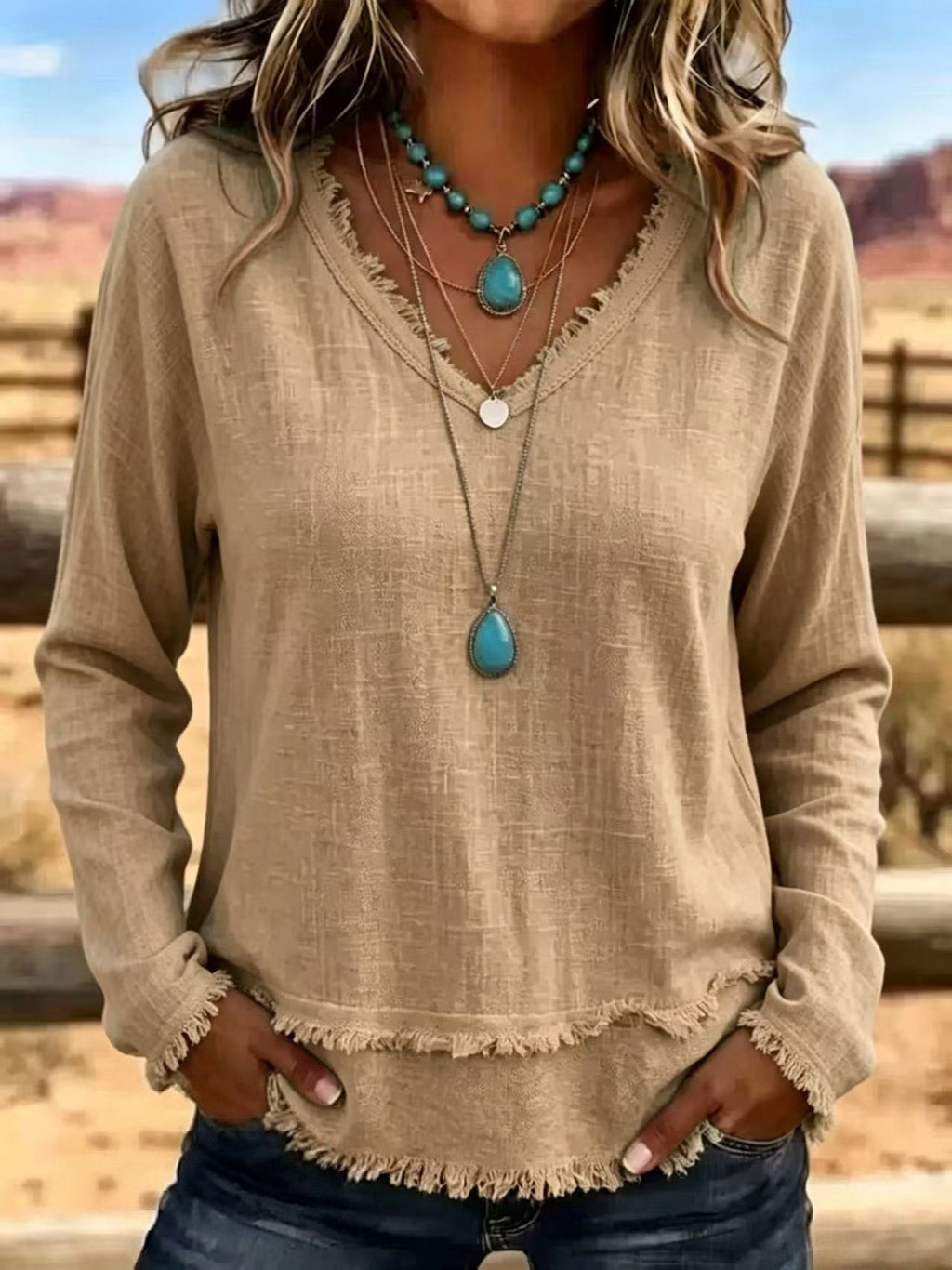 A woman wearing a beige long-sleeve V-neck top with a frayed hem and cuffs, layered with turquoise and silver necklaces, set against a blurred outdoor desert background.