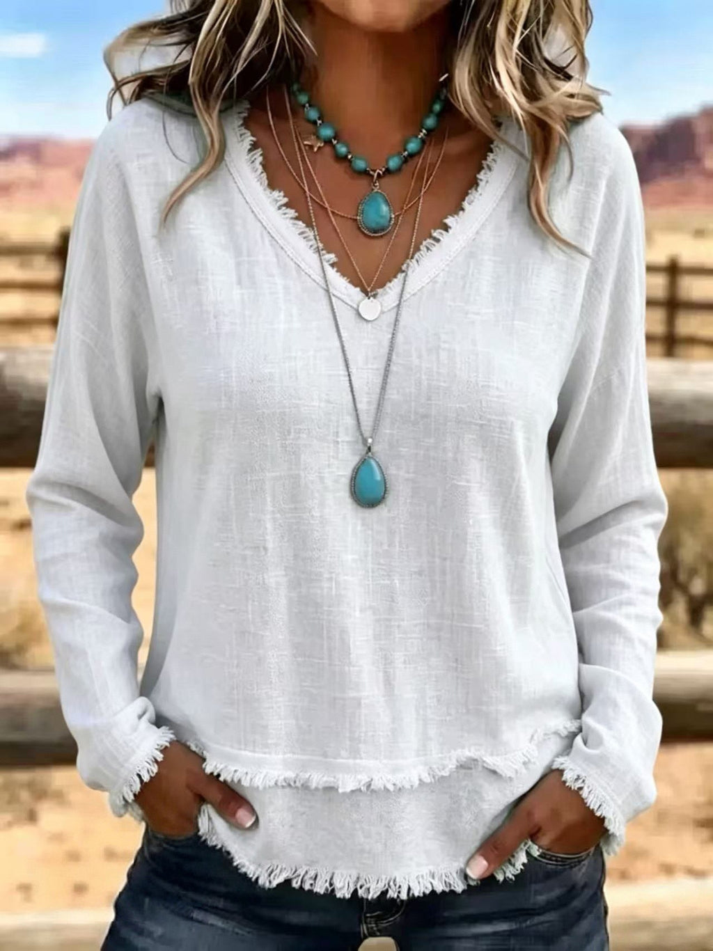 A woman wearing a white long-sleeve linen top with a frayed hem and cuffs, layered with turquoise and silver necklaces, standing outdoors in a desert landscape.