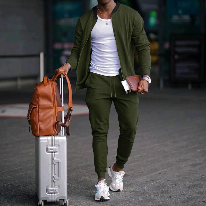 Man in green jacket and pants with a suitcase and brown backpack walking on a street.