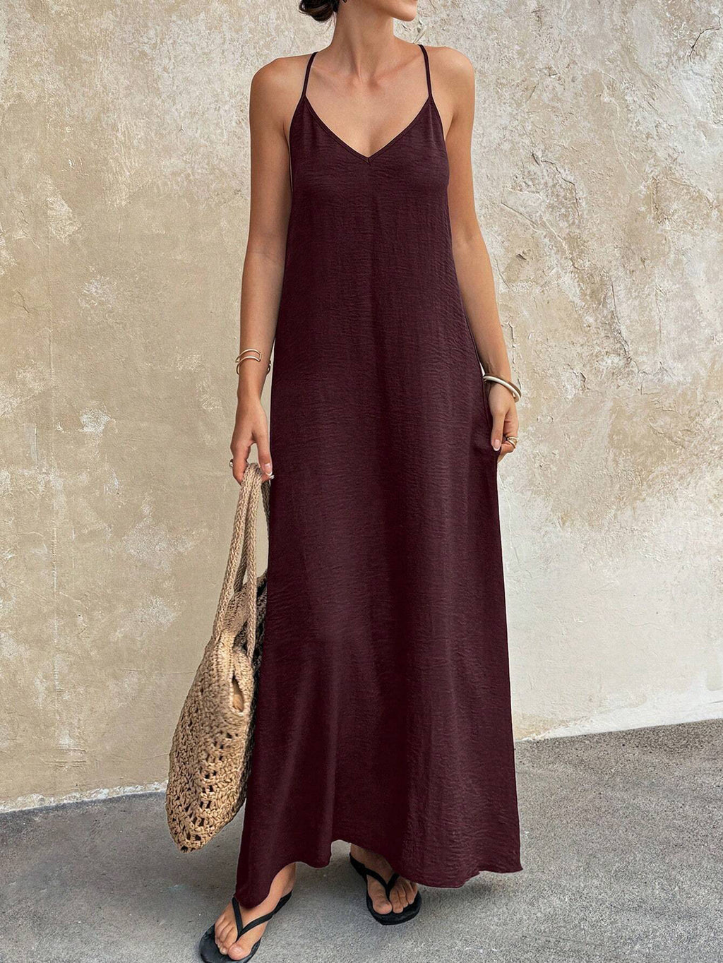 Woman wearing a long brown dress holding a woven bag against a textured wall.