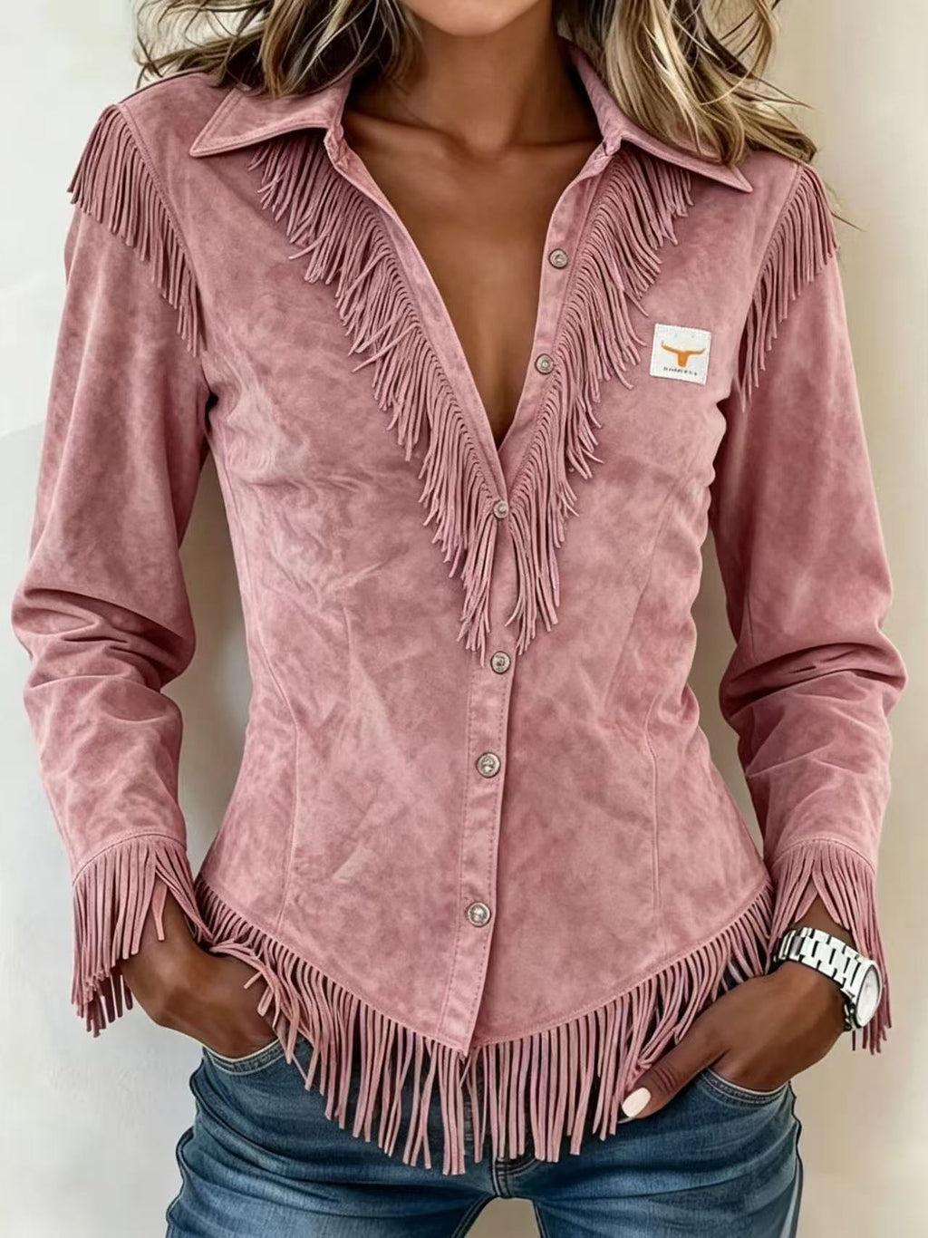 A woman wearing a dusty pink suede long-sleeve shirt with fringe detailing and a small Texas longhorn logo patch on the chest, paired with blue jeans.
