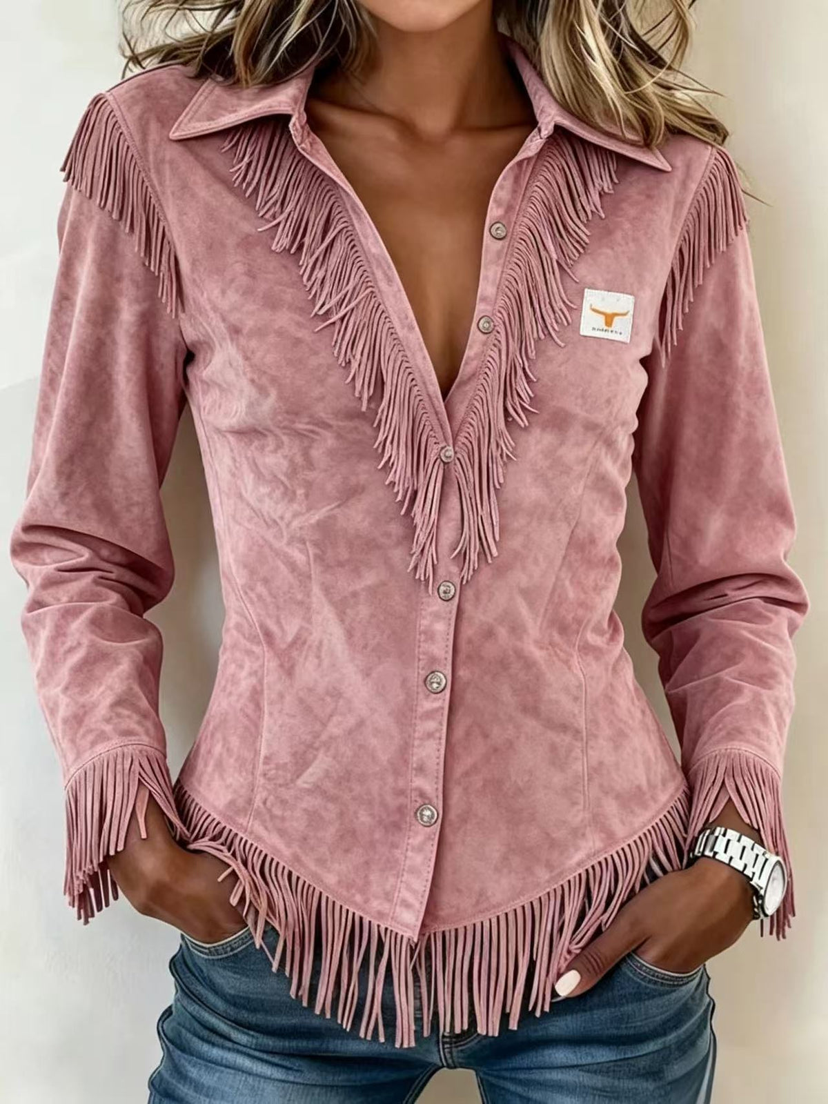 A woman wearing a dusty pink suede long-sleeve shirt with fringe detailing and a small Texas longhorn logo patch on the chest, paired with blue jeans.