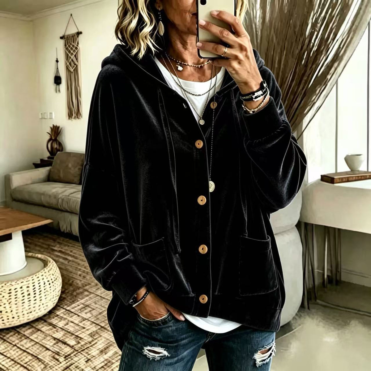 A woman takes a mirror selfie wearing a black velvet button-up hoodie jacket with brown buttons, layered over a white t-shirt and blue ripped jeans in a modern living room setting.