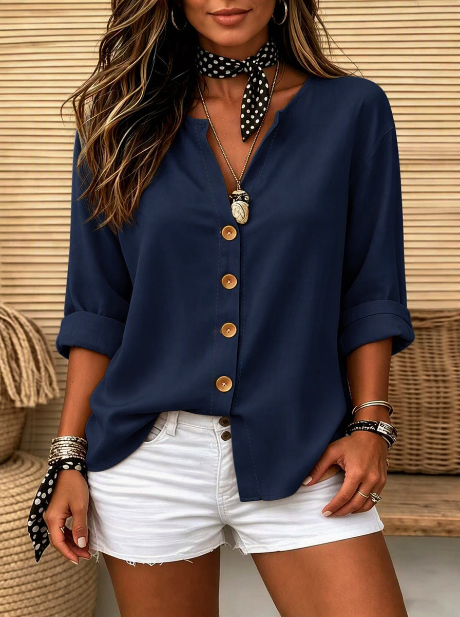 A woman wearing a navy blue button-up blouse with rolled sleeves and white denim shorts, styled with a black-and-white polka dot neck scarf and layered necklaces, set against a wicker and wood slat background.