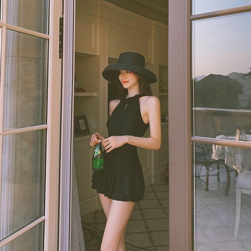 Woman in a black dress and hat standing by a glass door, holding a green bottle.