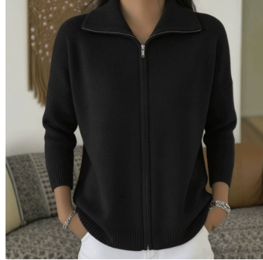 A person wearing a black ribbed zip-front cardigan sweater with a stand collar, paired with white bottoms, standing indoors in front of a sofa.