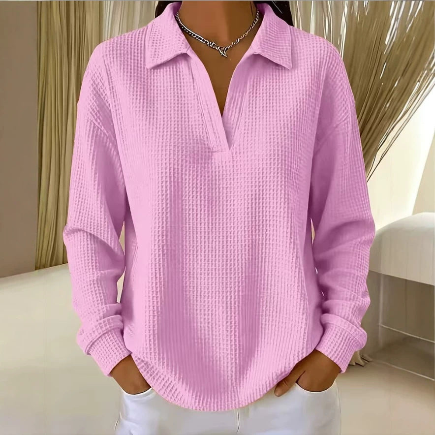 A woman wearing a light pink waffle-knit long-sleeve polo shirt with a split V-neck, paired with white pants and a silver chain necklace, standing in an indoor setting with neutral-toned curtains.