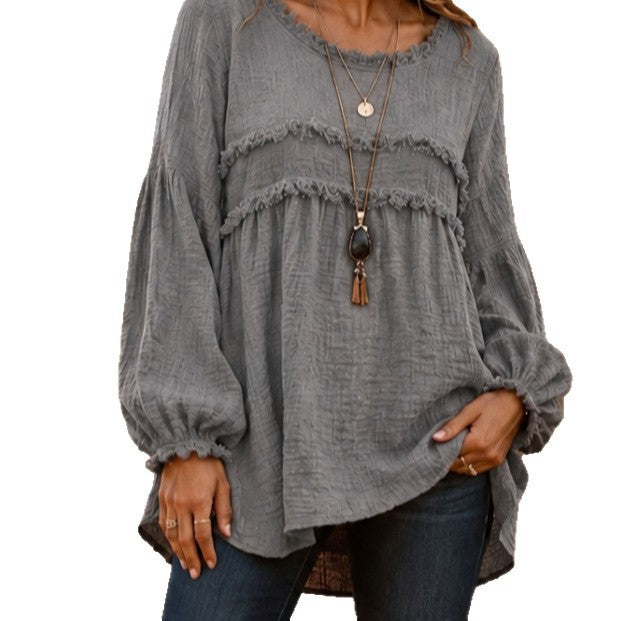 A woman wearing a gray long-sleeve blouse with a textured crinkle fabric, layered ruffle details across the chest, and a relaxed silhouette, paired with dark blue jeans and a long necklace.